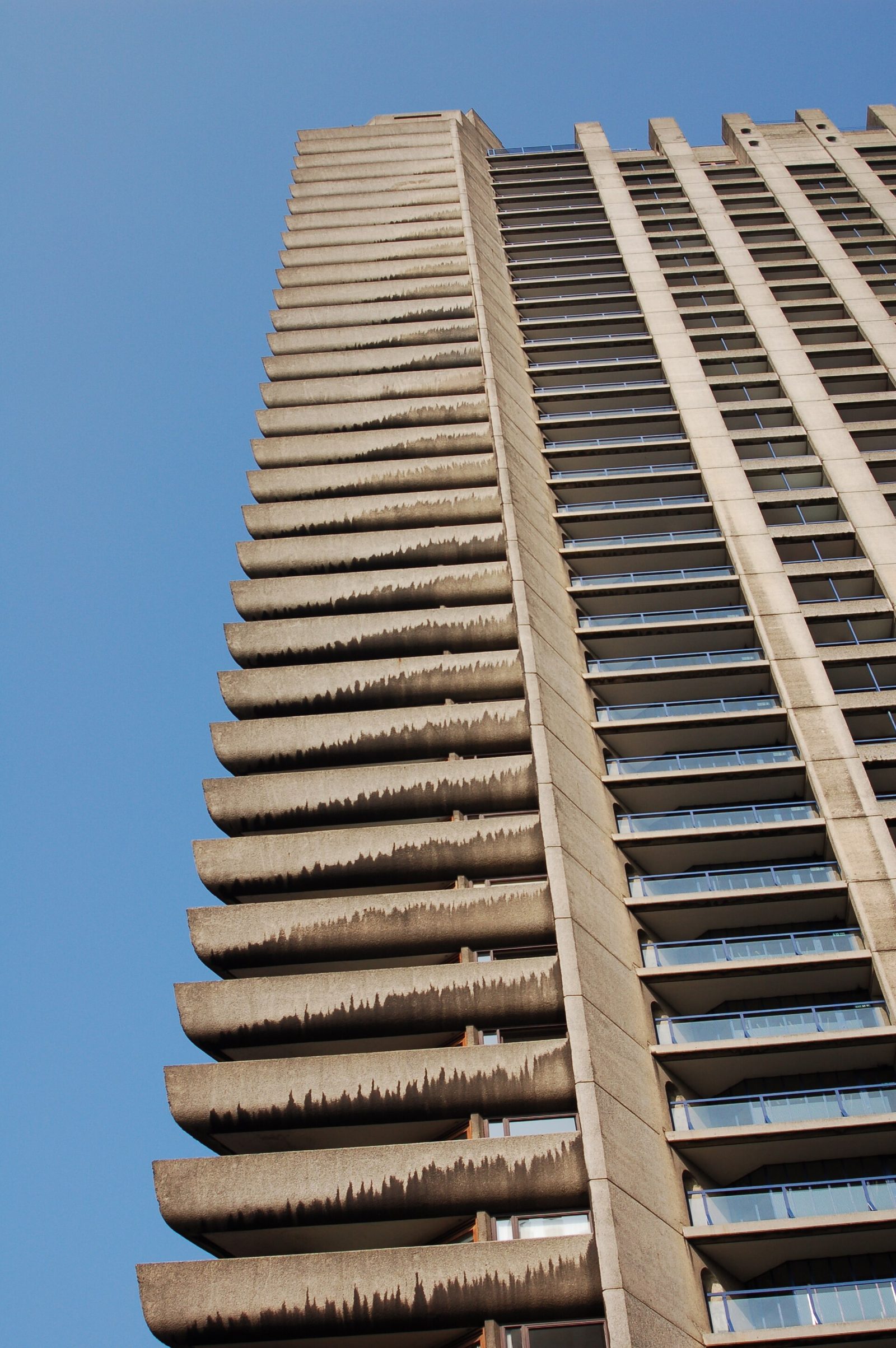 Jedna z wież Barbican Estate w Londynie (ukończono 1976, architekci Chamberlin, Powell and Bon) — beton bez okładziny po czterdziestu latach użytkowania: naturalne przebarwienia, spłukiwanie kruszyw, ślady deszczu wzdłuż spoin. To jest béton brut w eksploatacji — materiał wchodzi w czas. Fot. Todd Huffman, CC BY 2.0, Wikimedia Commons