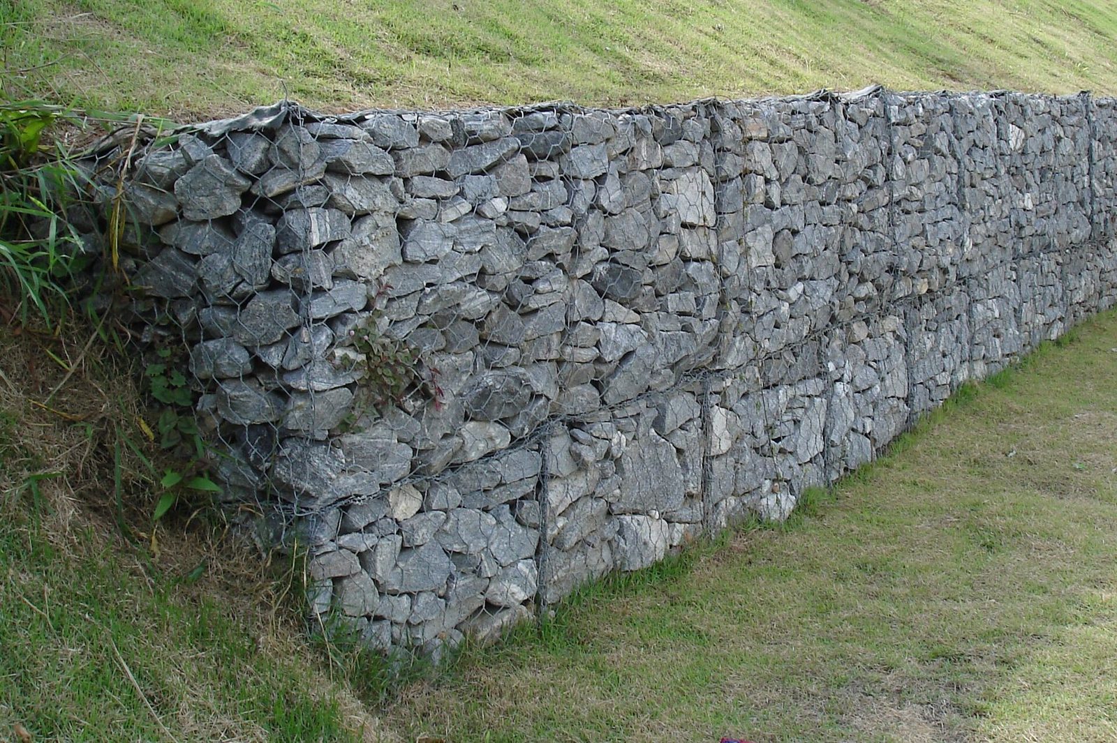 Mur gabionowy — stalowe kosze siatkowe wypełnione łamanym kamieniem, ułożone w terasy przy zielonym zboczu. Nieregularne kamienie wypełniają siatkę bez zaprawy. Fot. Zimbres, CC BY-SA 2.5, Wikimedia Commons